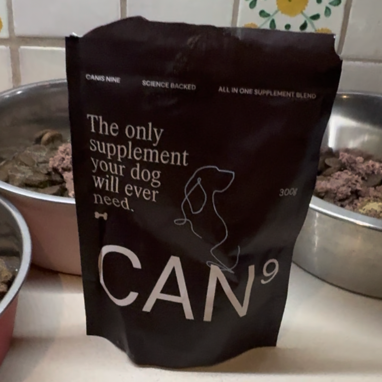 Black supplement bag labeled 'CAN9' on a tiled kitchen counter with pet food bowls in the background.